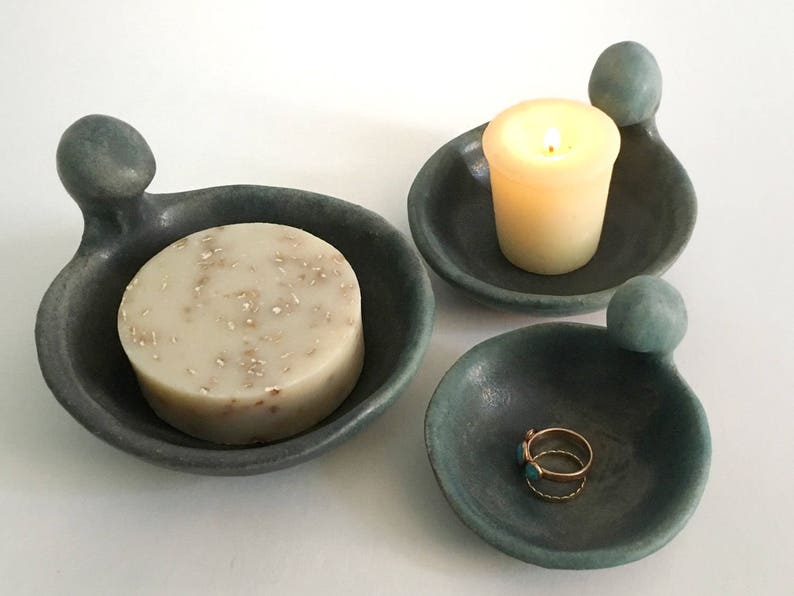 May include: Three ceramic dishes with a unique, organic shape. The dishes are in shades of teal and grey. One dish holds a bar of soap, another holds a candle, and the third holds a ring.