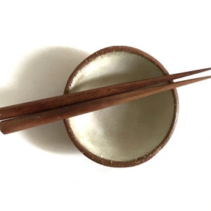 May include: A small, white ceramic bowl with a brown rim, resting on a white surface. Two wooden chopsticks are placed on top of the bowl.