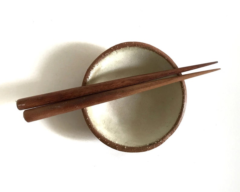 May include: A small, white ceramic bowl with a brown rim, resting on a white surface. Two wooden chopsticks are placed on top of the bowl.