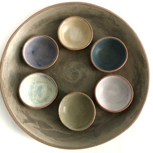 May include: A set of six small ceramic bowls in various colors, including blue, green, purple, and white, arranged in a circle on a larger, gray ceramic plate.