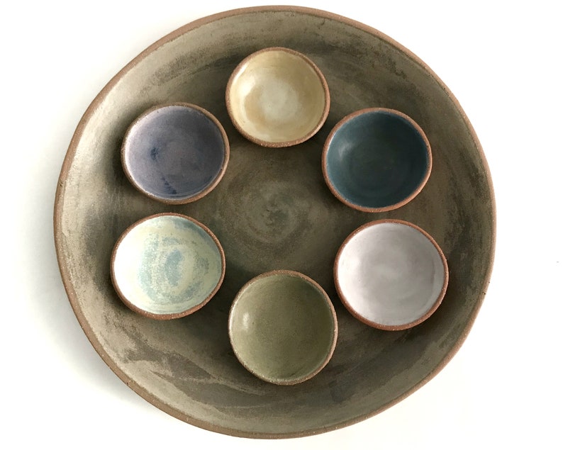 May include: A set of six small ceramic bowls in various colors, including blue, green, purple, and white, arranged in a circle on a larger, gray ceramic plate.