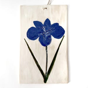 May include: A rectangular ceramic wall plaque with an off-white background featuring a hand-painted blue iris flower with green stems and leaves. The piece has a small hole at the top center for hanging, showcasing a glossy, textured finish.