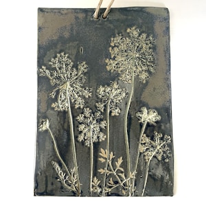 May include: A dark gray ceramic tile with a pressed design of white wildflowers. The tile has a rustic, aged look and is perfect for adding a touch of nature to your home decor.