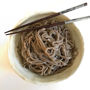 May include: A white bowl filled with brown soba noodles, with two wooden chopsticks resting on the rim of the bowl.