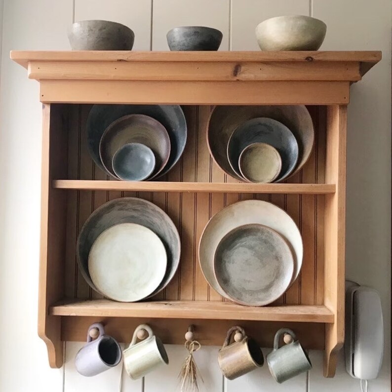May include: A wooden shelf unit with two shelves, displaying a collection of ceramic bowls and plates in various shades of brown, gray, and white. The shelves are mounted on a white wall, and there are four hooks underneath the shelves, each holding a ceramic mug.