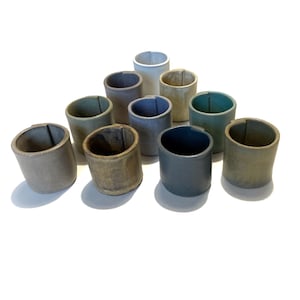 May include: A collection of ten ceramic cups in various shades of brown, green, and gray. The cups are arranged in a pyramid shape on a white surface.