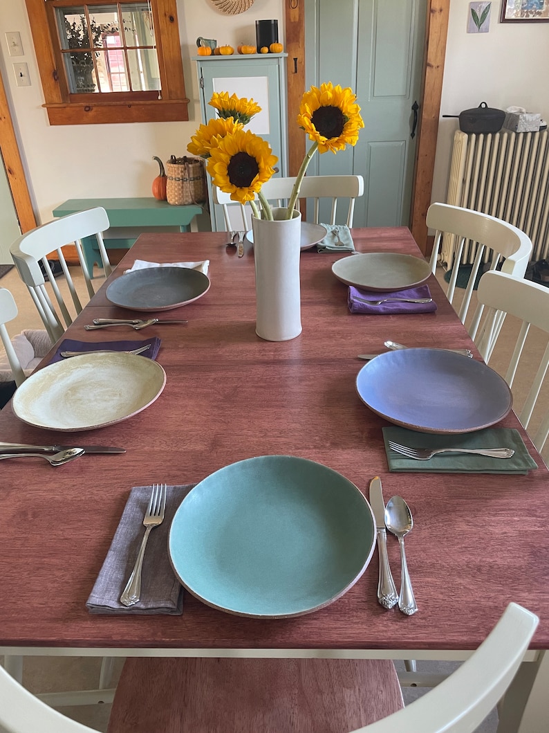 Puede incluir: Una mesa de comedor para seis personas con platos de cer&aacute;mica coloridos, cubiertos y servilletas de lino. Un jarr&oacute;n blanco contiene girasoles. La mesa es de madera rojiza y las sillas est&aacute;n pintadas de blanco.