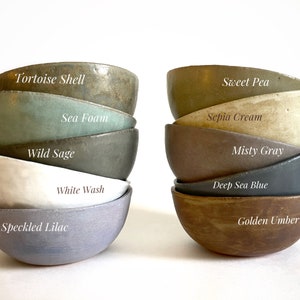 May include: A stack of ten small ceramic bowls in various shades of green, blue, brown, and gray. The bowls are labeled with names like "Tortoise Shell", "Sea Foam", "Wild Sage", "White Wash", "Speckled Lilac", "Sweet Pea", "Sepia Cream", "Misty Gray", "Deep Sea Blue", and "Golden Umber".