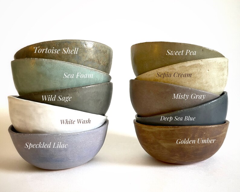 May include: A stack of ten small ceramic bowls in various shades of green, blue, brown, and gray. The bowls are labeled with names like "Tortoise Shell", "Sea Foam", "Wild Sage", "White Wash", "Speckled Lilac", "Sweet Pea", "Sepia Cream", "Misty Gray", "Deep Sea Blue", and "Golden Umber".