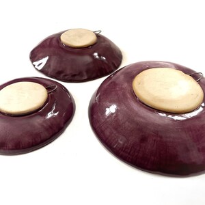 May include: Three purple ceramic plates with a wooden backing and a metal hanger on the back. The plates are different sizes.