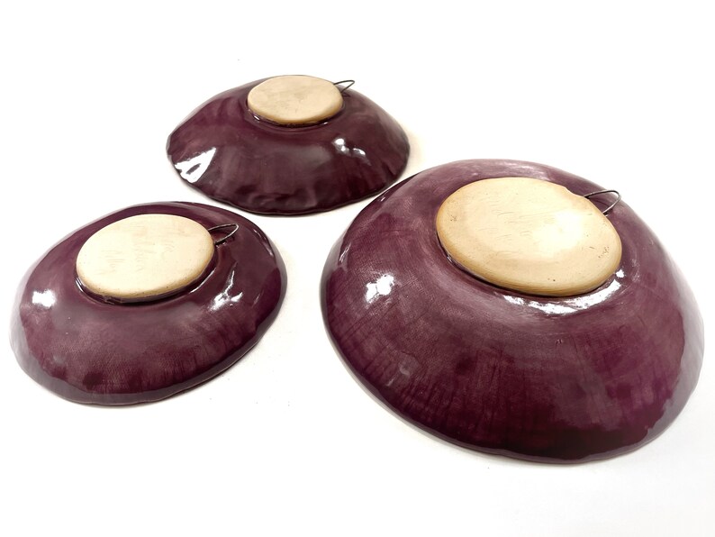 May include: Three purple ceramic plates with a wooden backing and a metal hanger on the back. The plates are different sizes.