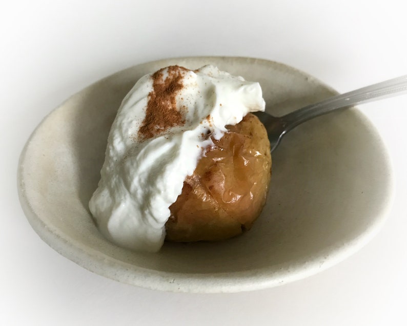 Puede incluir: Una manzana asada cubierta con crema batida y espolvoreada con canela, servida en un cuenco peque&ntilde;o de cer&aacute;mica blanquecina. Una cuchara plateada est&aacute; en el cuenco, lista para disfrutar del postre. El postre est&aacute; sobre un fondo blanco.