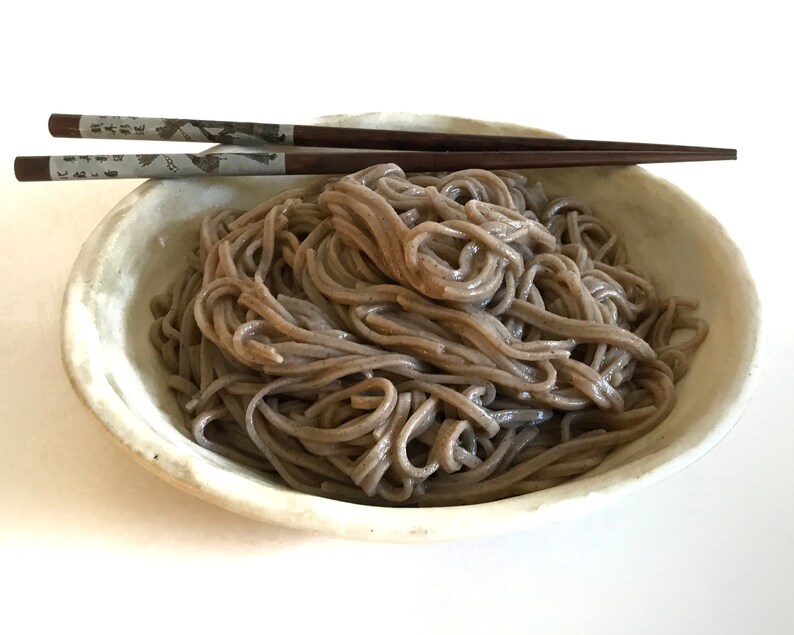 May include: A white bowl filled with a large portion of cooked soba noodles. The noodles are dark brown in color and are arranged in a loose pile. A pair of wooden chopsticks are resting on the rim of the bowl.