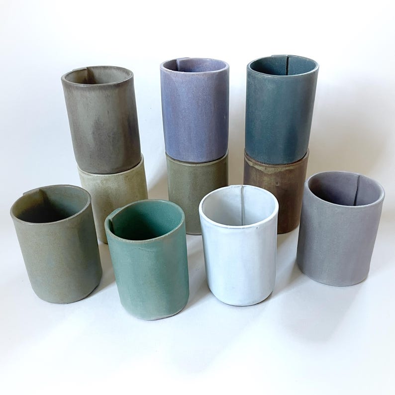 May include: Set of ten ceramic cups in various shades of green, brown, purple, and white. The cups are stacked in pairs, with the top cup of each pair being a different colour than the bottom cup.