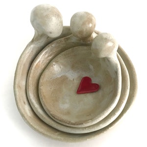 May include: Three ceramic bowls nested together, each shaped like a person with arms wrapped around the bowl below. The bowls are a light beige colour with a white glaze. A small red heart is in the centre of the smallest bowl.