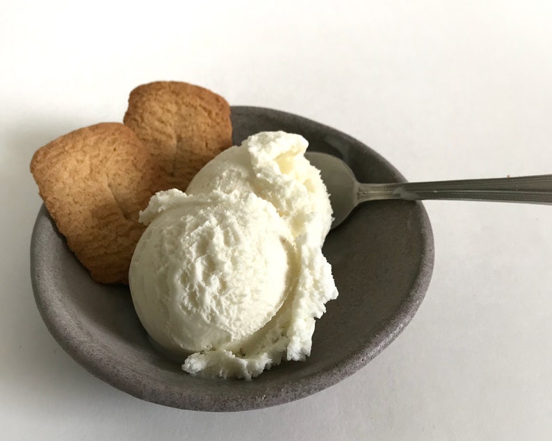 Puede incluir: Una porci&oacute;n de helado de vainilla con dos galletas doradas en un cuenco de cer&aacute;mica gris. Una cuchara plateada descansa en el borde del cuenco. El helado tiene un aspecto suave y texturizado.