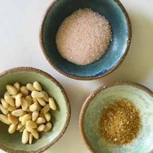 May include: Three small ceramic bowls with different toppings. The bowl on the left contains pine nuts, the middle bowl contains pink Himalayan salt, and the bowl on the right contains a yellow spice.