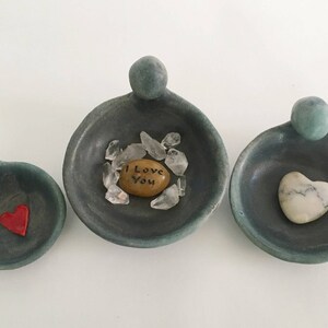 May include: Three small, teal-coloured ceramic dishes with rounded edges and a raised lip. The dishes are shaped like a person with a rounded head and a small body. Each dish contains a different object: a heart-shaped stone, a heart-shaped piece of red glass, and a small, round stone with the words "I Love You" written on it. The dishes are arranged in a row from left to right.