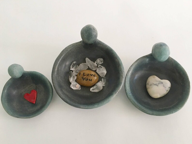 May include: Three small, teal-coloured ceramic dishes with rounded edges and a raised lip. The dishes are shaped like a person with a rounded head and a small body. Each dish contains a different object: a heart-shaped stone, a heart-shaped piece of red glass, and a small, round stone with the words "I Love You" written on it. The dishes are arranged in a row from left to right.