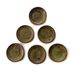 May include: Six small, round, olive-green ceramic dishes arranged in a triangular pattern. Each dish has a slightly raised rim with a darker, brownish hue. The dishes are set against a clean, white background.