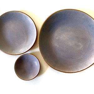 May include: Three ceramic bowls in various sizes, all with a light purple glaze and a brown rim. The bowls are arranged in a staggered formation on a white background.