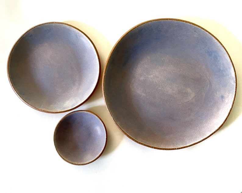 May include: Three ceramic bowls in various sizes, all with a light purple glaze and a brown rim. The bowls are arranged in a staggered formation on a white background.