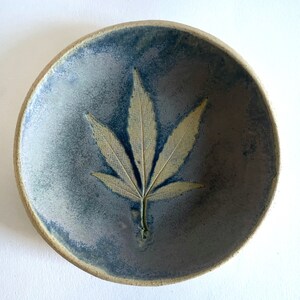 May include: A ceramic bowl with a blue and purple glaze features a pressed leaf design in the center. The leaf is a light yellow color and resembles a cannabis leaf.