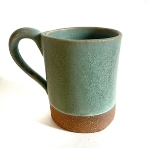 May include: A green ceramic mug with a brown rim and a speckled glaze. The mug has a rounded handle.