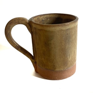 May include: A brown ceramic mug with a rounded handle. The mug has a light brown glaze with a darker brown rim and base.