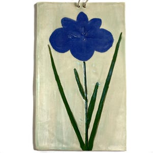 Puede incluir: Un colgante de pared de cerámica con una flor azul y tallos verdes sobre un fondo blanco.