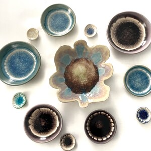May include: Assortment of small ceramic dishes in various shades of blue, green, and brown with a speckled glaze. The dishes are arranged in a circle on a white background.