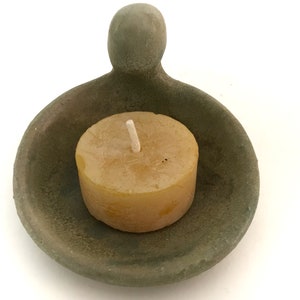 May include: A small, green ceramic candle holder with a rounded, human-shaped handle. A small, round, yellow candle sits inside the holder.
