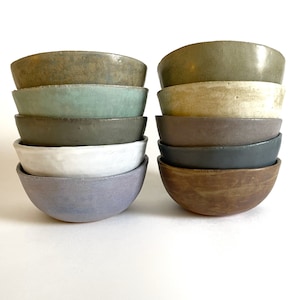 May include: Two stacks of ceramic bowls in various colors, including green, brown, white, and blue. The bowls have a simple, rounded design and a matte finish. The bowls are stacked on a white surface.
