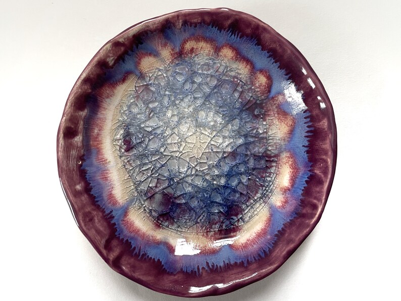 May include: A ceramic plate with a crackled glaze in shades of purple, blue, and white. The plate has a scalloped edge and a unique, organic design.
