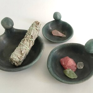May include: Three small, teal-coloured ceramic dishes with rounded edges and a raised knob on one side. The dishes are filled with various items, including a bundle of sage, a pink crystal, and a smaller clear crystal.