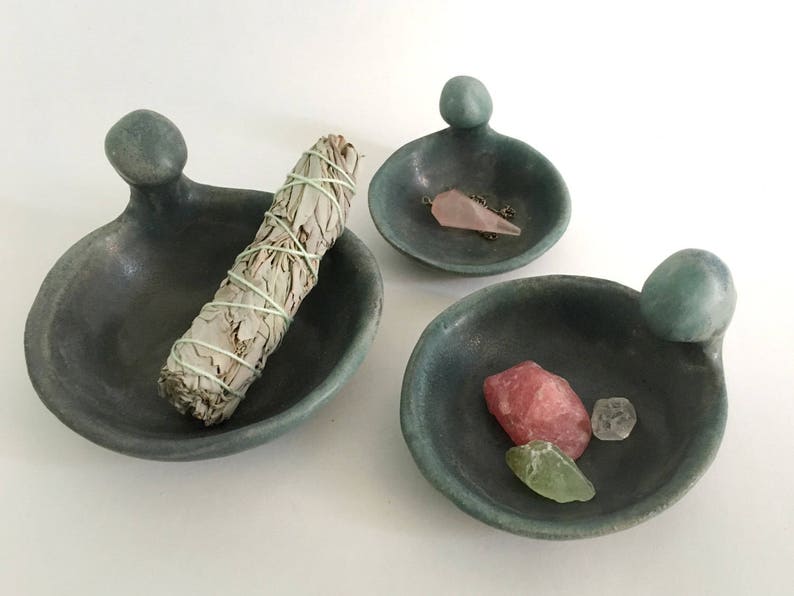 May include: Three small, teal-coloured ceramic dishes with rounded edges and a raised knob on one side. The dishes are filled with various items, including a bundle of sage, a pink crystal, and a smaller clear crystal.