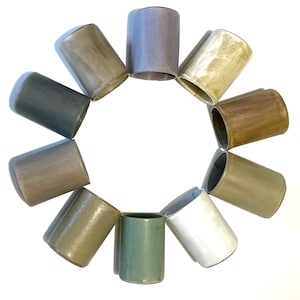 May include: A set of 10 ceramic cups in various shades of brown, grey, and green. The cups are arranged in a circle on a white background.