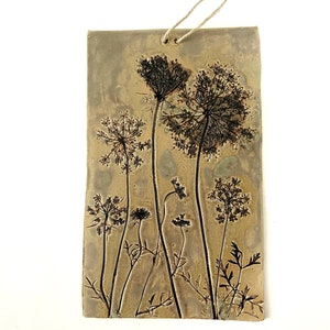 May include: A rectangular ceramic wall hanging with a rustic brown background and a pressed design of black dried wildflowers. The piece has a hole at the top for hanging.