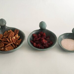 May include: Three small, teal-coloured ceramic bowls with handles shaped like human figures. The bowls are filled with pecans, dried cranberries, and white sugar.