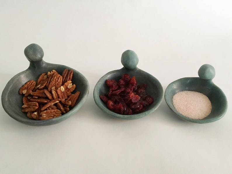 May include: Three small, teal-coloured ceramic bowls with handles shaped like human figures. The bowls are filled with pecans, dried cranberries, and white sugar.