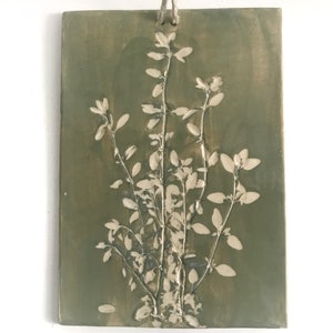Könnte beinhalten: Eine rechteckige Wandplatte aus Keramik in gedämpftem Olivgrün mit einem erhabenen, cremeweißen botanischen Relief einer blättrigen Pflanze. Das Stück hat eine glatte, glänzende Oberfläche und ein Loch an der Oberseite zur Aufhängung mit einer Schnur.