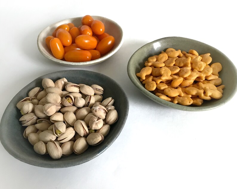 Puede incluir: Tres cuencos de cer&aacute;mica peque&ntilde;os e irregulares llenos de comida. Un cuenco contiene tomates cherry naranjas, otro contiene galletas de pez dorado y el tercero est&aacute; lleno de pistachos. Los cuencos son de tonos grises y beige.
