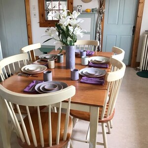 May include: A wooden dining table set with white chairs, a bouquet of white flowers in a blue vase, and a table setting with plates, bowls, and cups.