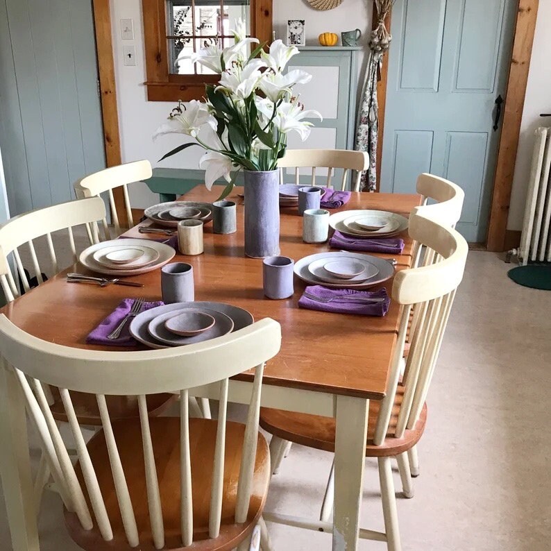May include: A wooden dining table set with white chairs, a bouquet of white flowers in a blue vase, and a table setting with plates, bowls, and cups.