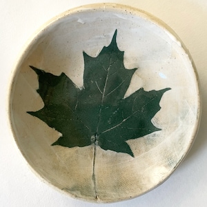 May include: A white ceramic bowl with a green maple leaf design. The bowl is perfect for serving snacks or holding small items.