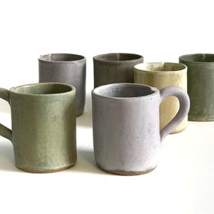 May include: Six ceramic mugs in various shades of green, purple, and beige. The mugs have a simple, rounded design and are perfect for everyday use.