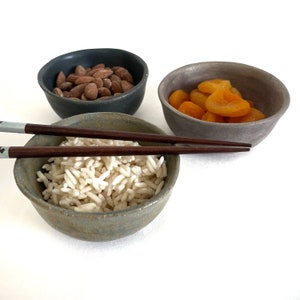 May include: Three small ceramic bowls, one gray, one blue-gray, and one green-gray, filled with almonds, dried apricots, and white rice. A pair of wooden chopsticks are resting on the rim of the bowl with rice.