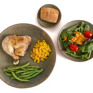 May include: A green ceramic plate with a roasted chicken leg, green beans, and corn kernels. A smaller green ceramic plate with a salad of green lettuce, shredded carrots, and cherry tomatoes. A small brown ceramic dish with a round bread roll.