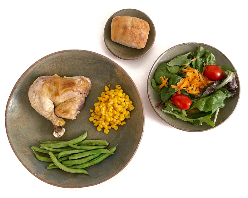 May include: A green ceramic plate with a roasted chicken leg, green beans, and corn kernels. A smaller green ceramic plate with a salad of green lettuce, shredded carrots, and cherry tomatoes. A small brown ceramic dish with a round bread roll.