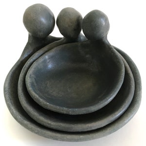 May include: Three grey ceramic nesting bowls with rounded handles. The bowls are stacked on top of each other.